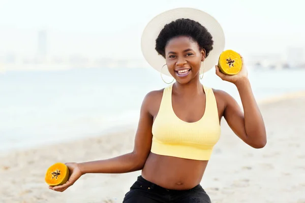 Uma mulher africana bonita em um chapéu de palha e com um mamão suculento em suas mãos está sentada na areia na praia, sorrindo e olhando para a câmera — Fotografia de Stock