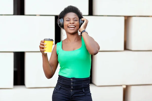 Mulher africana sorridente segura café em uma xícara descartável, ouve uma melodia agradável em fones de ouvido e danças — Fotografia de Stock
