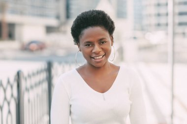 Beyaz kazaklı genç bir Afrikalı kadın gülümsüyor ve şehirde dolaşıyor. Yumuşak odak arkaplanı, metin için boşluk