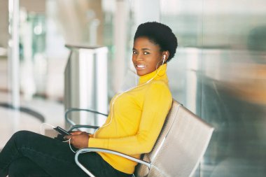 Güzel Afrikalı kadın bekleme odasında bir sandalyede oturmuş metro trenini bekliyor ve kulaklıkla müzik dinliyor.