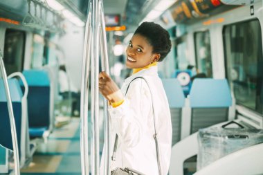 Gülümseyen genç bir Afrikalı kadın işe giderken metroda dururken tırabzana tutunuyor.