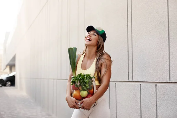 Uma menina esportiva sorri e segura um saco cheio de frutas e legumes frescos. Estilo de vida saudável, saúde, beleza, esportes — Fotografia de Stock