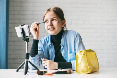Genç bir kız, blogu için evdeki bir tripoda akıllı telefondan video kaydederken kaşlarını ve kirpiklerini nasıl boyayacağını gösteriyor. arkadaşlar