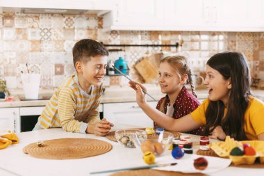 İki kız kardeş ve bir erkek kardeş masada oturmuş Paskalya için farklı renklerde yumurta boyuyor, gülüyor ve şımartıyorlar.