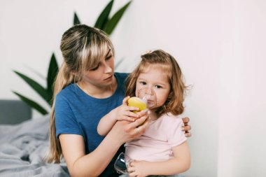 Anne hasta bir kızın nebulizör kullanmasına yardım ediyor. Kanepede ona sarılıyor. Anne hasta bir çocuğun nefes almasına yardım ediyor.