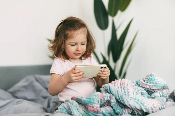A little girl watches cartoons on her phone or plays on the bed before going to bed