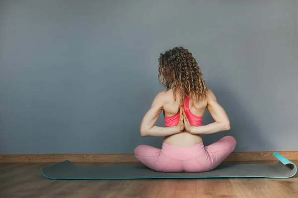 Uma menina com cabelo encaracolado senta-se em uma posição de lótus em um tapete de ioga e medita depois de um treino. Visão traseira, espaço para texto — Fotografia de Stock