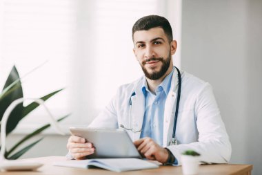 Doğu görünümlü sevimli genç bir doktor masada oturur ve kameraya bakarak dostça gülümser..
