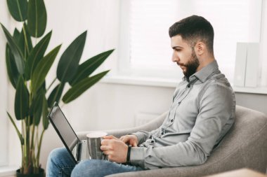 Doğulu görünümlü yakışıklı genç adam evde çalışıyor, dizüstü bilgisayarlı bir sandalyede oturuyor.