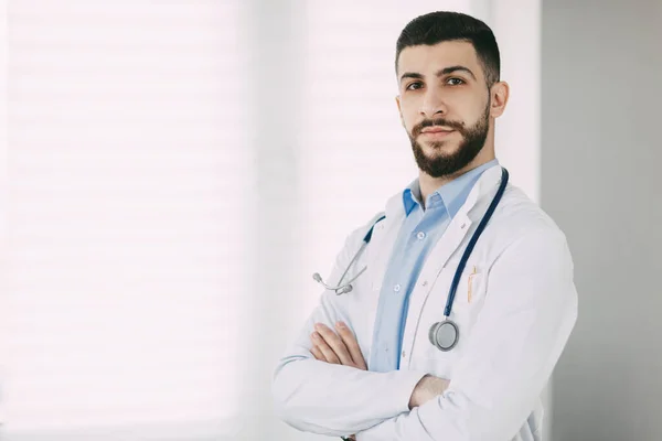 Poważny młody lekarz w stetoskopie i białym płaszczu medycznym stoi w swoim biurze z skrzyżowanymi ramionami — Zdjęcie stockowe