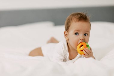 Çocuk kar beyazı bir yatakta yatıyor, gülüyor ve çıngırakla oynuyor..