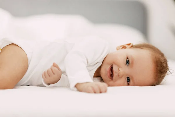En blåögd bebis ligger i en snövit säng under en filt, skrattar och njuter — Stockfoto