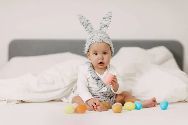 赤ちゃんはウサギの衣装でベッドの上に座って、カラフルなイースターエッグで遊んで、笑い、楽しいです — ストック写真