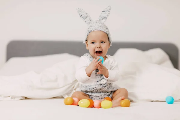 赤ちゃんはウサギの衣装でベッドの上に座って、カラフルなイースターエッグで遊んで、笑い、楽しいです — ストック写真