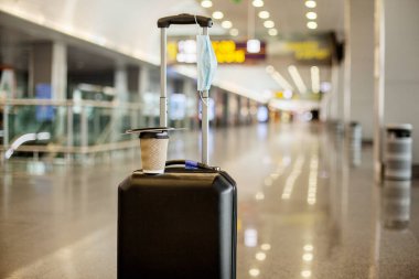 Boş bir havaalanında, bir bavul, bir bardak kahve ve tıbbi koruyucu bir maske vardır. Coronavirus, sosyal uzaklık, izolasyon