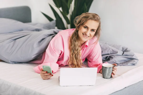 Uma linda garota está deitada na cama com uma camisa rosa, segurando uma xícara de café na mão e usando um laptop para ver conteúdo online, treinamento on-line, trabalho on-line — Fotografia de Stock