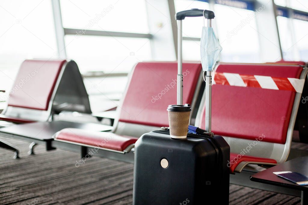 Next to the seats in an empty airport is a suitcase, on it is a glass of coffee, hanging a medical protective mask. Coronavirus, social distance, isolation