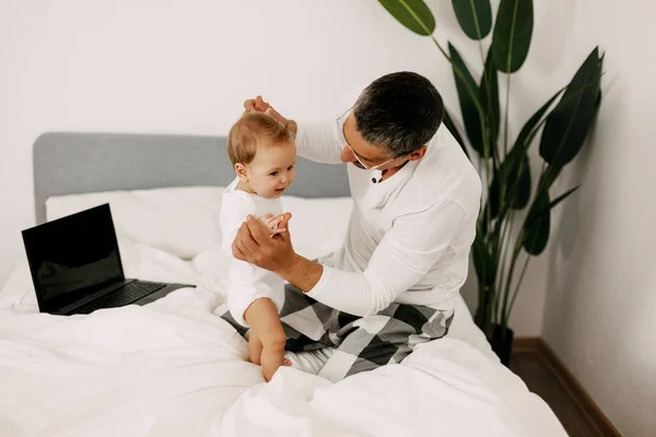 Dad sits on the bed, works on a laptop at home with a baby on his lap, smiles and looks at the screen together — Stock Photo, Image