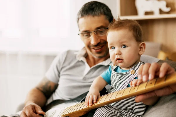 A middle-aged father with tattoos on his arms sits in an armchair in the room and plays the guitar to his little one.