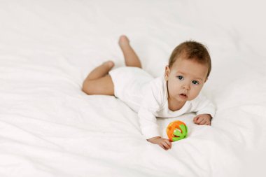 Çocuk kar beyazı bir yatakta yatıyor, gülüyor ve çıngırakla oynuyor..