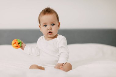 Çocuk kar beyazı bir yatakta oturuyor, gülüyor ve çıngırakla oynuyor..