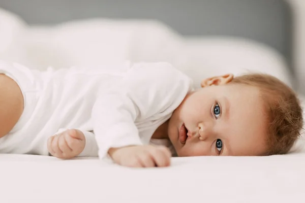 En blåögd bebis ligger i en snövit säng under en filt, skrattar och njuter — Stockfoto