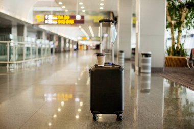 Boş bir havaalanında, bir bavul, bir bardak kahve ve tıbbi koruyucu bir maske vardır. Coronavirus, sosyal uzaklık, izolasyon