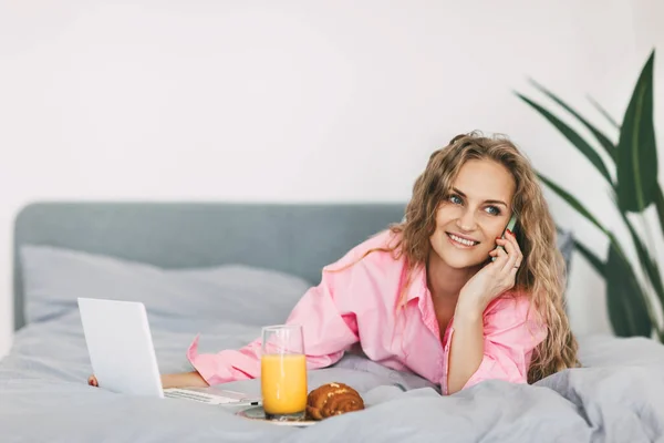 Uma jovem está ao telefone, deitada na cama durante o café da manhã, com um laptop ao lado dela. Tecnologias modernas, freelancer, comunicação online — Fotografia de Stock