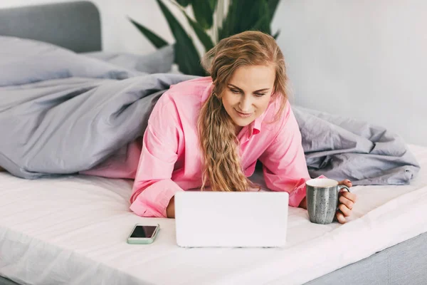 한 아름다운 소녀가 분홍색 셔츠를 입은 채 침대에 누워 손에 커피를 들고 컴퓨터를 사용하여 온라인 콘텐츠를 보고 온라인 교육을 받고 온라인으로 일하고 있습니다. — 스톡 사진