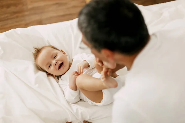 Glad far leker med bedårande bebis i sovrummet i en snövit säng — Stockfoto