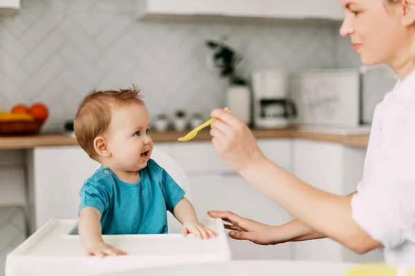 Genç bir anne, yüksek bir bebek beslenme sandalyesinde oturan küçük oğlunu besler. Bakım, annelik, çocukluk