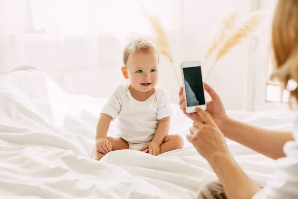 Bébé mignon est assis sur le lit et posant pour sa mère, maman prend des photos de son bébé sur son téléphone portable. Technologies modernes, espace pour le texte — Photo