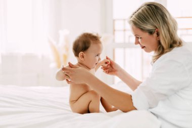 Sevimli bir bebek kollarını açmış ve annesinin ellerini tutarak yatakta oturuyor. Yeni beceriler, destek ve bakım geliştirin