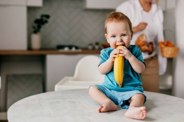 Küçük bir çocuk elinde muzla mutfak masasında oturuyor ve gülümsüyor.