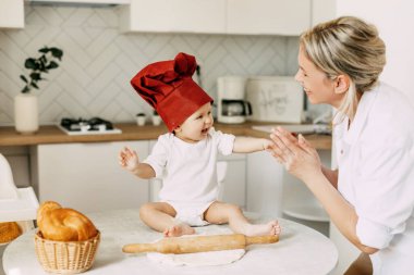Yemek pişirme şapkası takmış sevimli bir bebek masanın üzerinde oturur. Hamur, yemek pişirmek için yatar ve annesine gülümser.