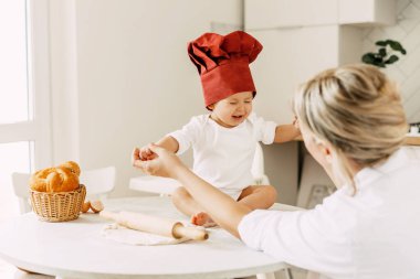 Yemek pişirme şapkası takmış sevimli bir bebek masanın üzerinde oturur. Hamur, yemek pişirmek için yatar ve annesine gülümser.