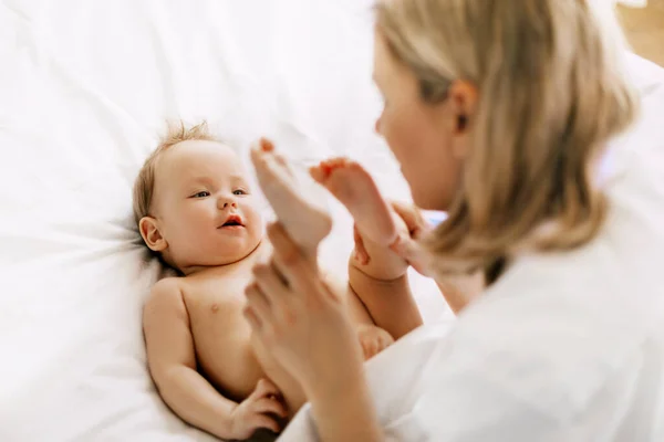 Felice mamma gioca con il suo bambino sul letto e tiene le gambe nelle sue mani — Foto Stock