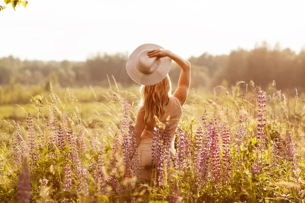 Uma jovem em um vestido leve e um chapéu em um campo de tremoço olha para o pôr do sol. Visão traseira, espaço para texto — Fotografia de Stock