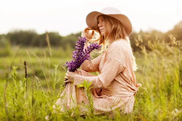 Portrait of a beautiful blonde with a bouquet of lupines in her hands sitting on the lawn and looking thoughtfully into the distance — Stock Photo, Image