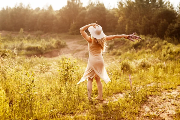 Uma jovem com um vestido leve e um chapéu com um buquê nas mãos está dançando no campo, olhando para o pôr do sol. Visão traseira, espaço para texto — Fotografia de Stock