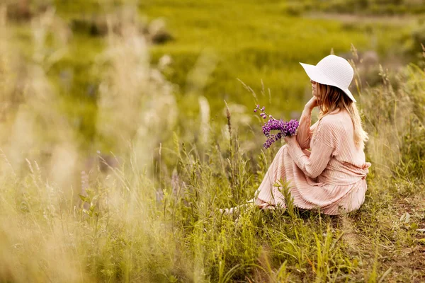 Retrato de uma bela loira com um buquê de tremoços em suas mãos sentado no gramado e olhando atentamente para a distância — Fotografia de Stock