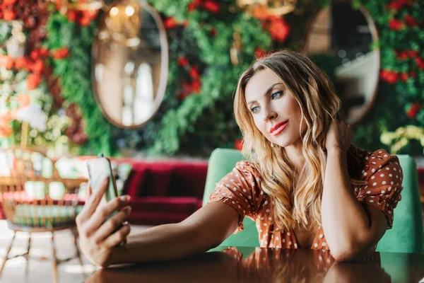 Una mujer joven toma una selfie en su teléfono y se sienta en una mesa en un hermoso café — Foto de Stock