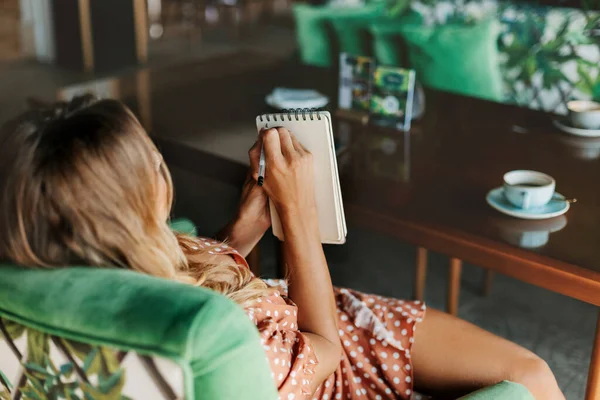 Uma menina loira está sentada em uma mesa em um café, bebendo café e fazendo anotações em seu caderno durante uma pausa — Fotografia de Stock