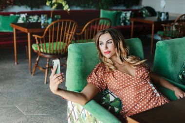 Genç bir kadın telefonuyla selfie çekiyor ve güzel bir kafedeki masada oturuyor.
