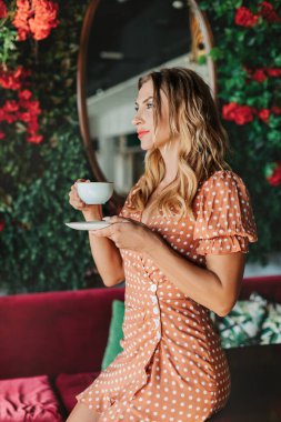 Genç bir kadın sabah bir restoranda kahve içer ve düşünceli bir şekilde uzaktan bakar, aromanın tadını çıkarır.