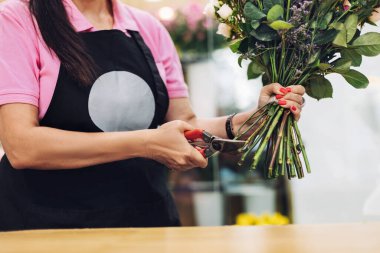Önlüklü profesyonel bir kadın çiçekçi masadaki çiçek sapını makasla keser. Çiçeklerle çalışma konsepti, çiçek işi..