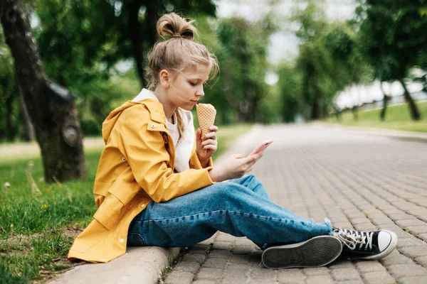 Blondýnka sedící na silnici s telefonem a zmrzlinou komunikuje na sociálních sítích. Dítě venku — Stock fotografie