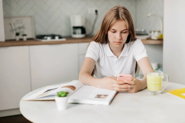 Uma adolescente está sentada em uma mesa na cozinha, segurando um telefone e fazendo trabalhos de casa — Fotografia de Stock