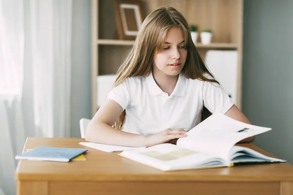 Una chica bonita está leyendo un libro sentado en una mesa, haciendo los deberes, preparándose para los exámenes. Educación, formación, conocimiento — Foto de Stock