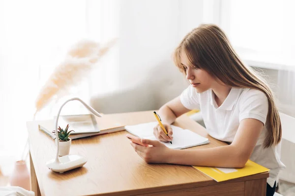 Una hermosa chica está sentada en una mesa y preparándose para los exámenes, sosteniendo un teléfono en sus manos, buscando información en Internet — Foto de Stock